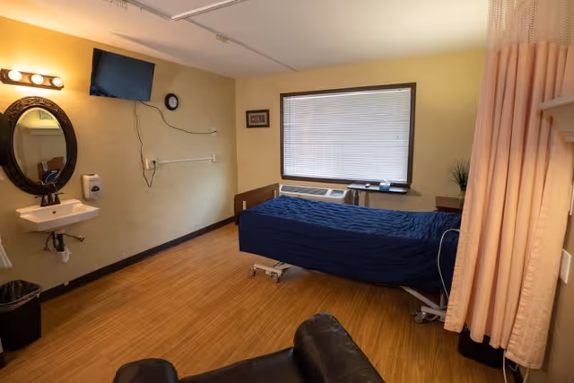A simple healthcare bedroom with a single bed covered in a blue blanket, wall-mounted TV, sink, window, and a privacy curtain.