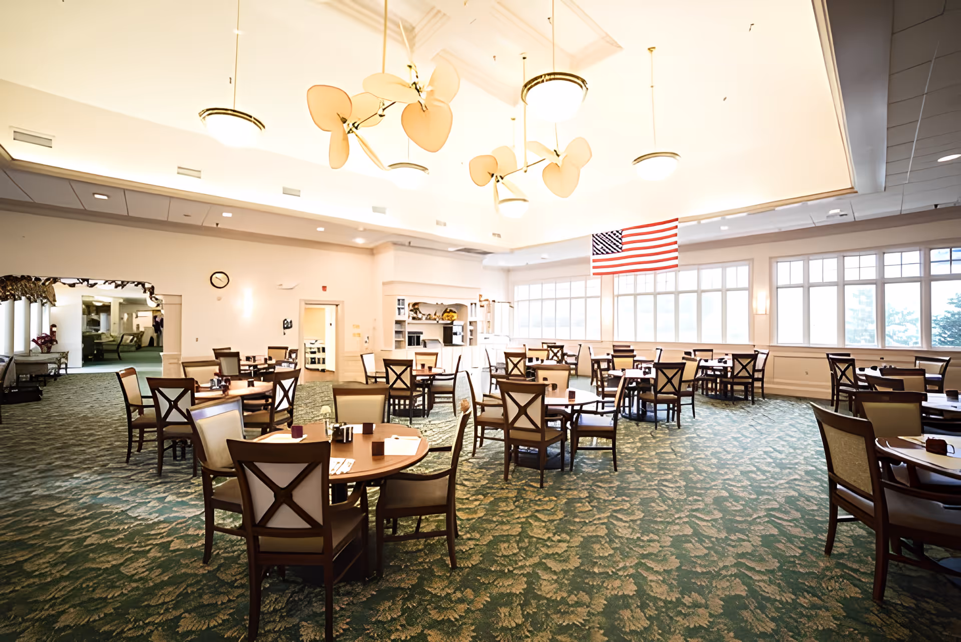 A spacious dining room with multiple round wooden tables and cushioned chairs arranged neatly. The room has large windows letting in natural light, an American flag hanging near the windows, and ceiling fans with light fixtures. The carpet has a green leafy pattern, and there is a doorway leading to another room.