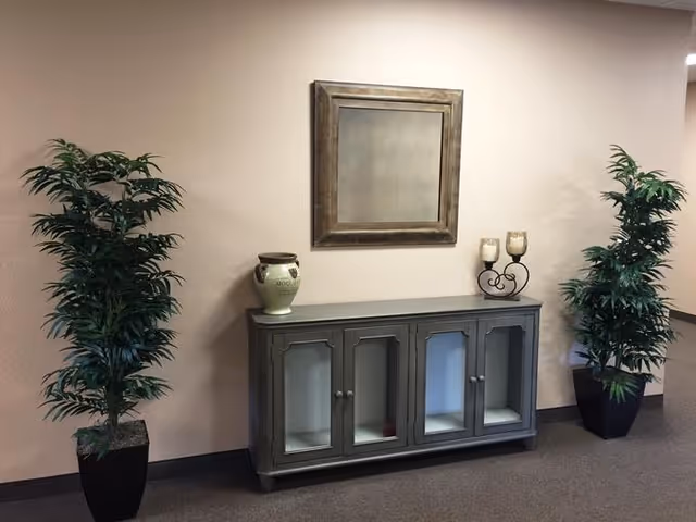 A decorative hallway vignette with a gray console cabinet, wall mirror, two potted plants and tabletop decor against a beige wall.