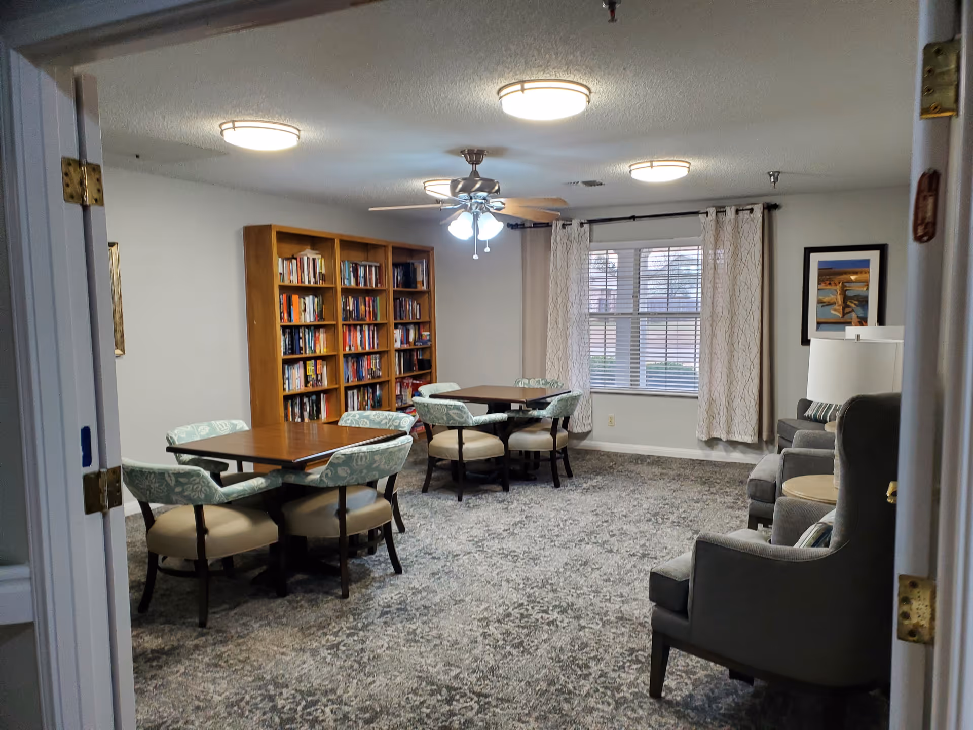 Communal sitting room with card tables and upholstered chairs, a large bookshelf, armchairs, and a window with curtains.
