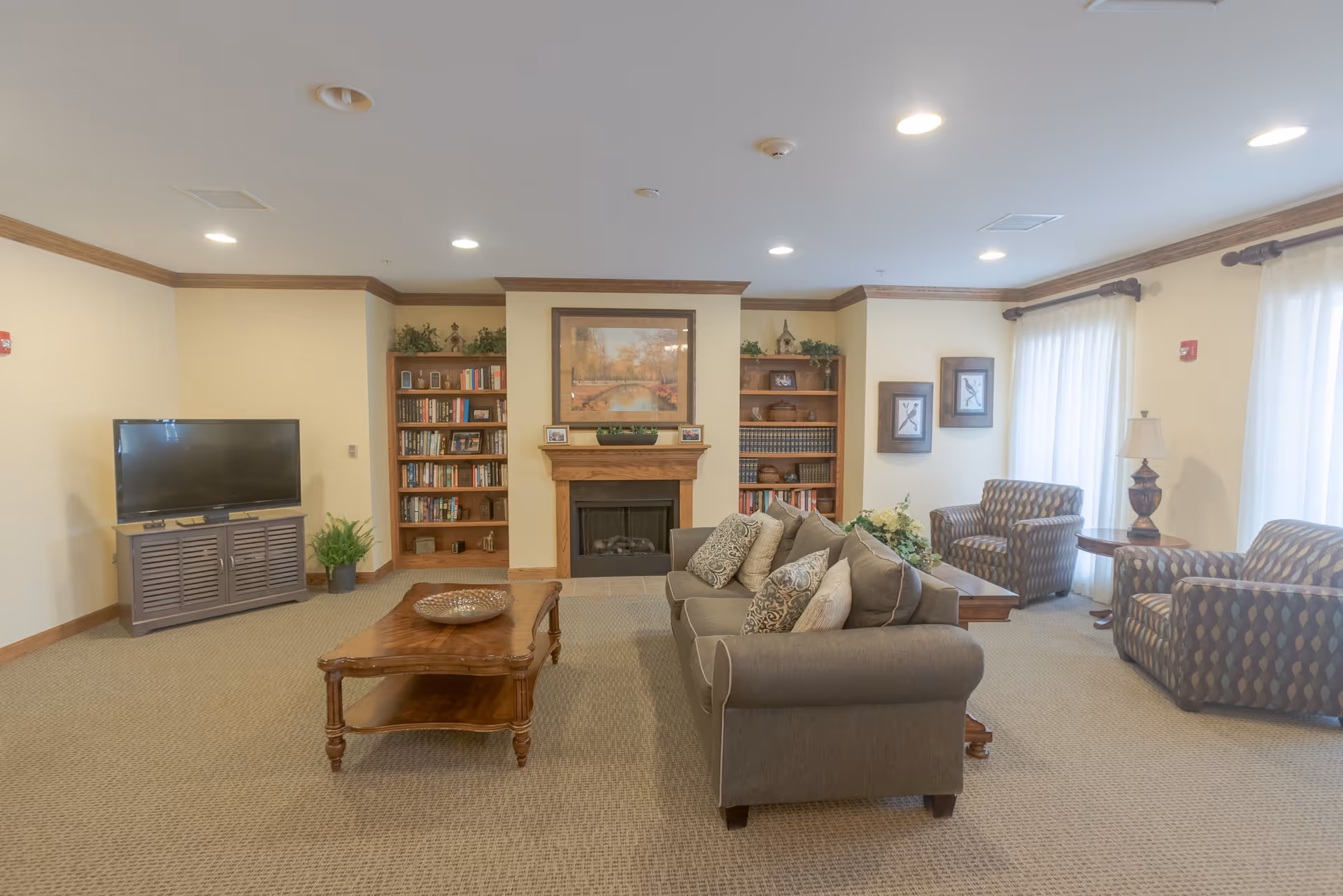 A cozy living room with a gray sofa adorned with patterned pillows, two patterned armchairs, a wooden coffee table, and a TV on a wooden stand. The room features a fireplace flanked by two bookshelves filled with books and decorative items. Light beige walls with crown molding and large windows with sheer white curtains allow natural light to fill the space.
