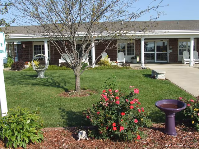 Front exterior view of a single-story brick building with a covered porch supported by white columns. The lawn in front has a tree, flowering bushes, a birdbath, and garden decorations. There are white chairs on the porch near the entrance.