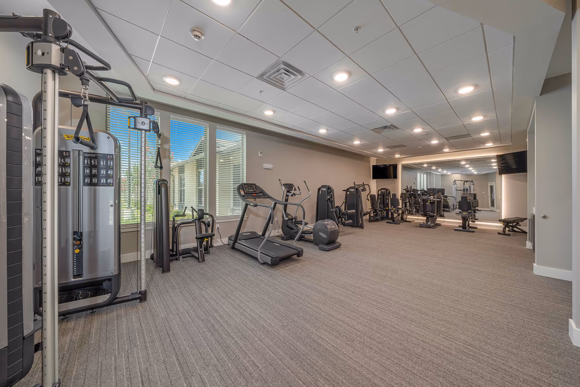 A fitness room with various exercise equipment including treadmills, elliptical machines, weight machines, and benches. The room has large windows with blinds, a carpeted floor, recessed ceiling lights, and a large mirror on one wall. Two flat-screen TVs are mounted on the walls.
