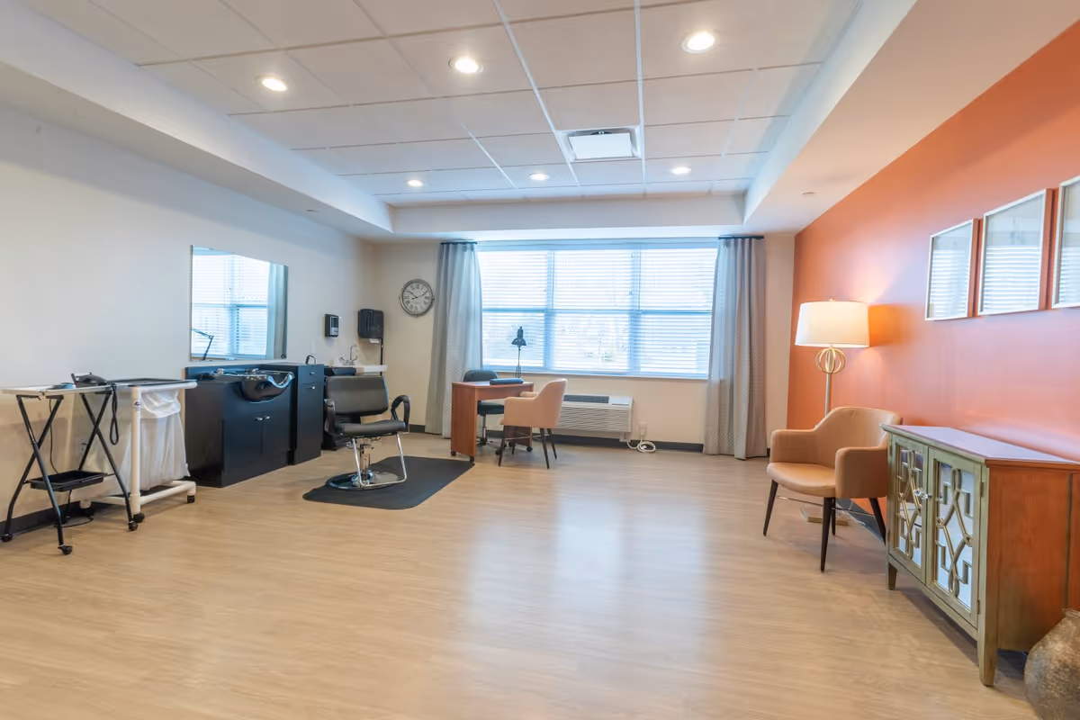 A spacious room with light wood flooring and a white ceiling with recessed lighting. On the left side, there is a black salon chair in front of a large mirror and a black sink station, along with a small rolling cart and a laundry hamper. In the center near the window, there is a small wooden desk with two beige chairs and a black desk lamp. The window has white blinds and gray curtains. On the right side, an orange accent wall features three framed mirrors above a wooden cabinet with mirrored doors, next to a beige armchair and a floor lamp with a white shade.