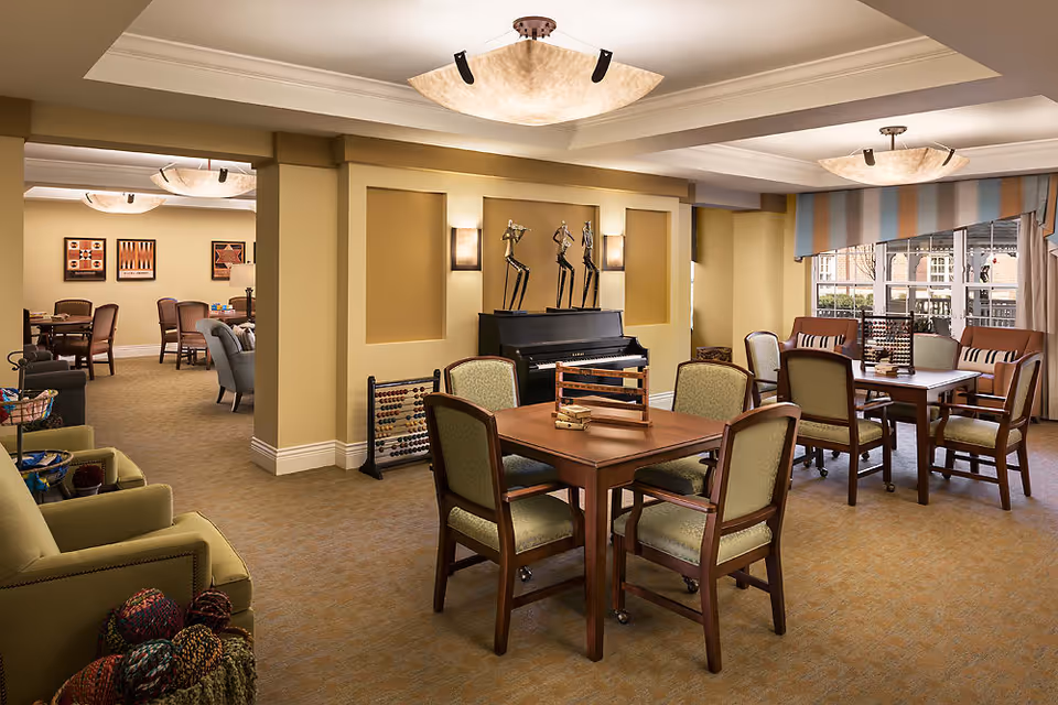 A spacious, well-lit common area in a senior living facility featuring multiple wooden tables with chairs arranged around them. The room has beige walls and carpet, decorative sculptures on a piano against the wall, and large windows with striped valances letting in natural light. There are also comfortable armchairs and framed artwork on the walls.