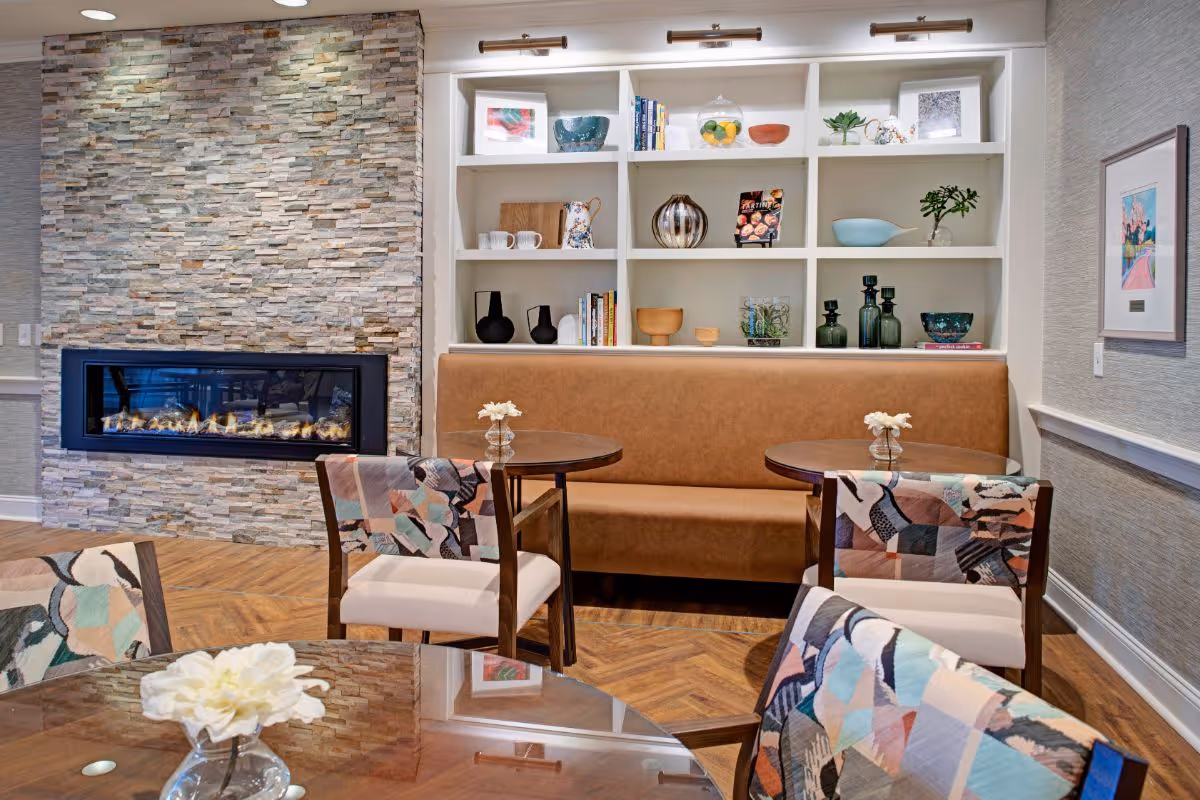 A cozy seating area in a senior living facility with a stone fireplace on the left, a built-in white shelving unit with decorative items and books, a brown cushioned bench, and round wooden tables with floral centerpieces. The chairs have colorful patterned upholstery, and there is a framed artwork on the wall to the right.