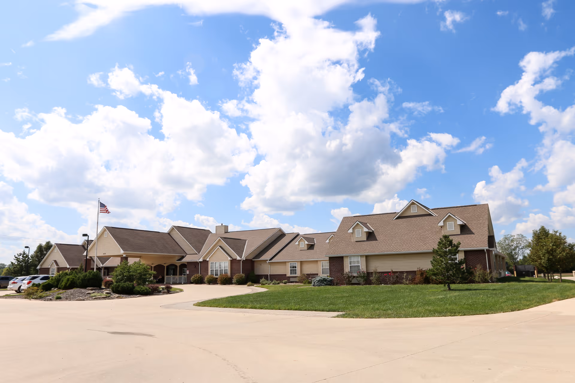 Exterior view of a single-story senior living facility building with a beige and brick facade, multiple gabled roofs, a flagpole with an American flag, a driveway, and a well-maintained lawn under a partly cloudy blue sky.