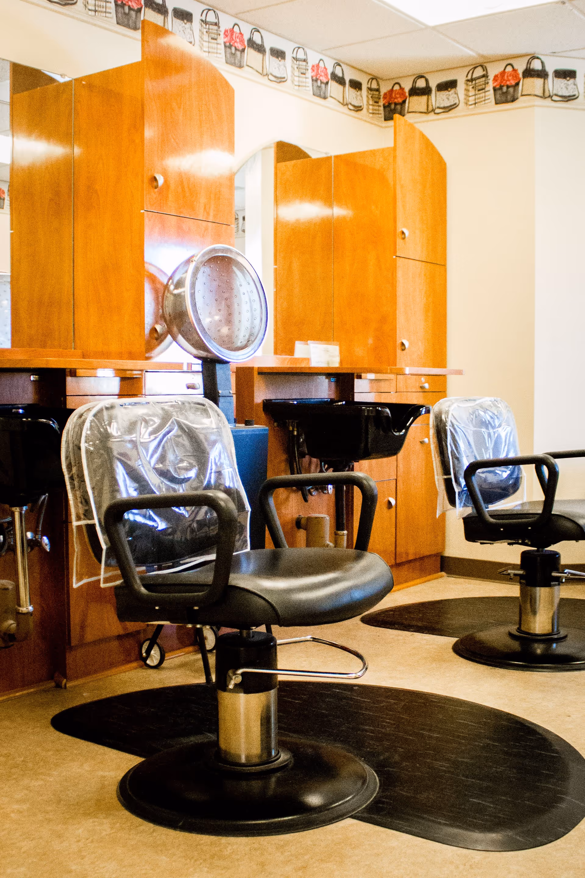 Interior view of a salon area with two black salon chairs covered with clear plastic, black sinks for hair washing, wooden cabinets with mirrors, and a decorative border featuring handbags along the top of the wall.