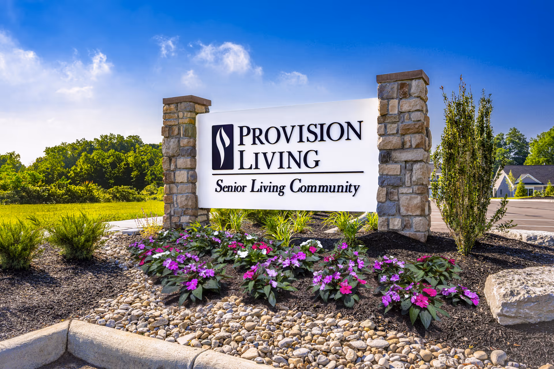 Entrance sign reading "Provision Living Senior Living Community" set between stone pillars with flowers and landscaping under a blue sky.