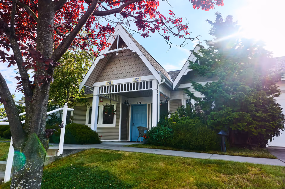 Front exterior of a cottage-style retirement building with a blue door, covered porch, and surrounding trees and lawn.