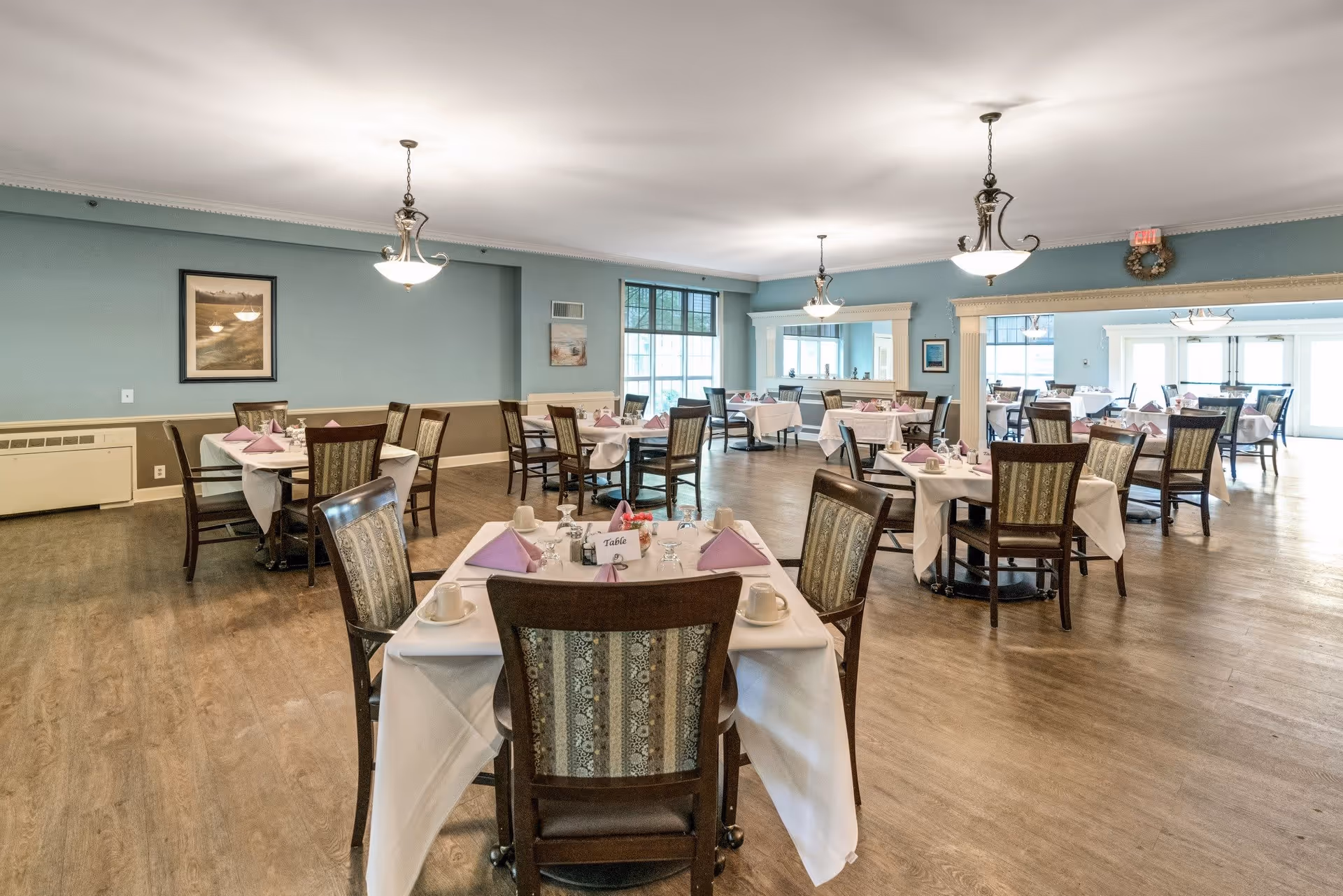 A spacious dining room with multiple tables covered in white tablecloths and set with cups, napkins, and utensils. The walls are painted light blue with framed artwork, and there are large windows allowing natural light to fill the room. The floor is wooden, and elegant light fixtures hang from the ceiling.