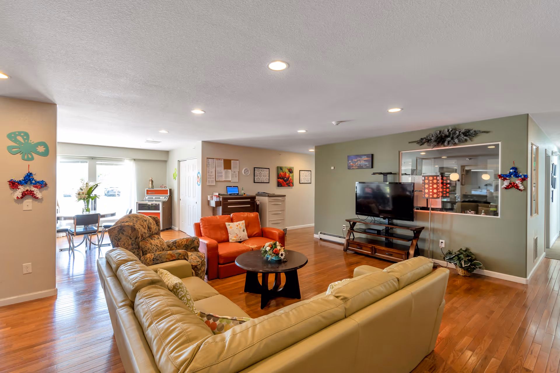 Bright communal living room with sofas, armchairs, a TV on a stand, coffee table and a dining area visible in the background.