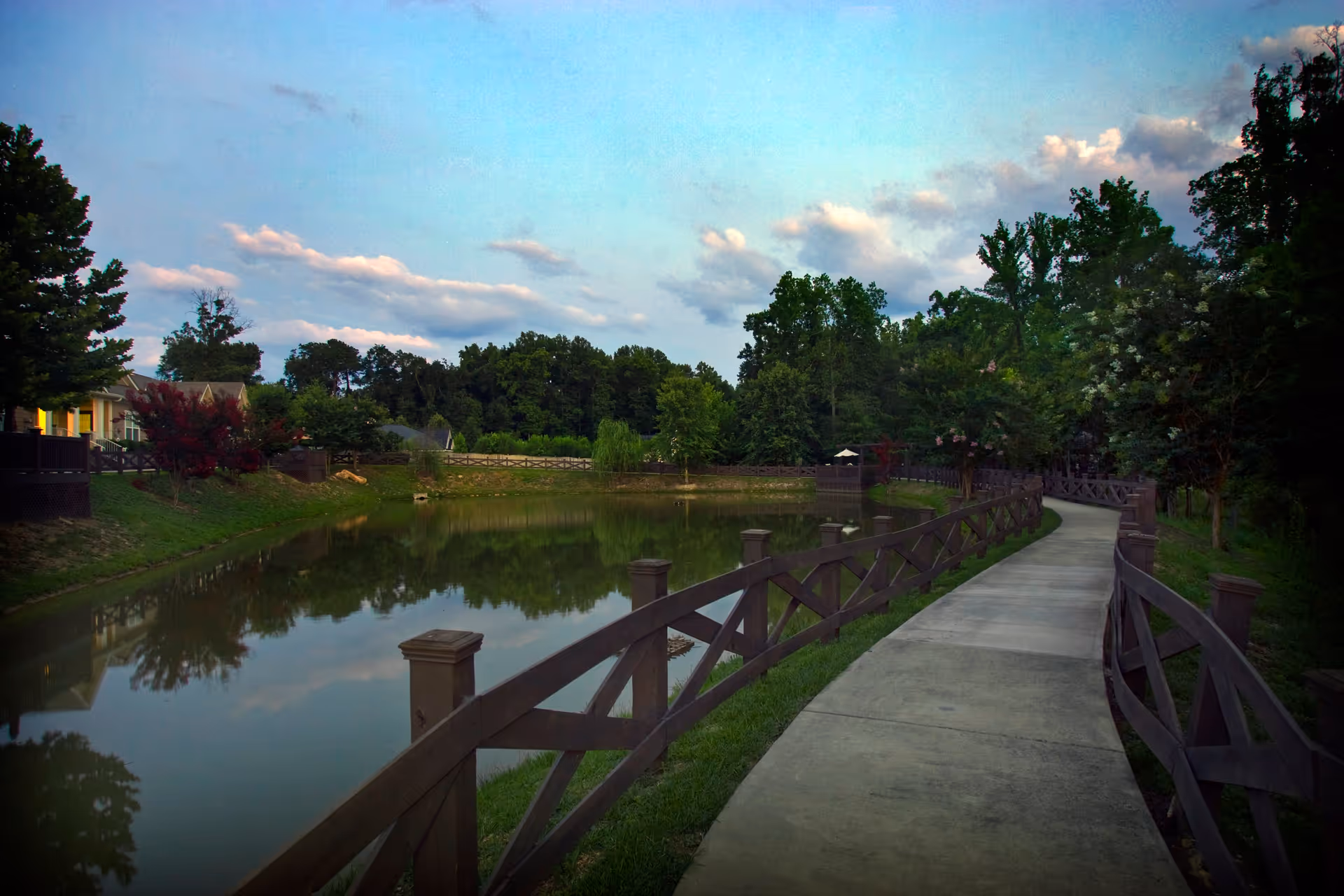 A winding concrete pathway bordered by wooden railings runs alongside a calm pond surrounded by green trees and shrubs under a partly cloudy sky.