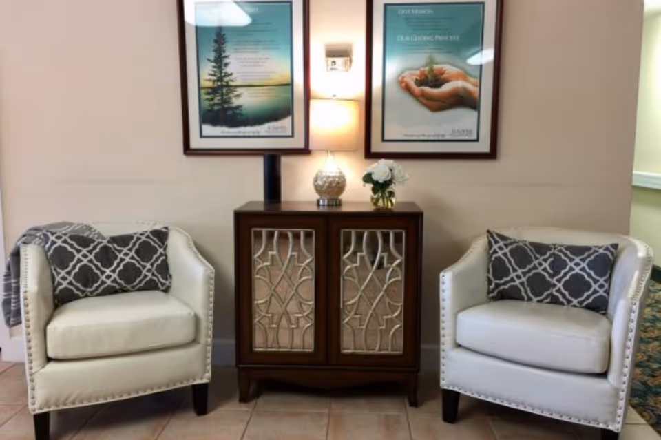 Two white upholstered armchairs flanking a wooden console table topped with a lamp and framed artwork on the wall.