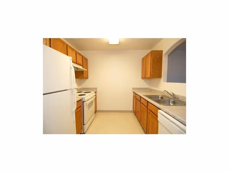 Small galley kitchen with white refrigerator, stove, dishwasher, double sink, and wooden cabinets.