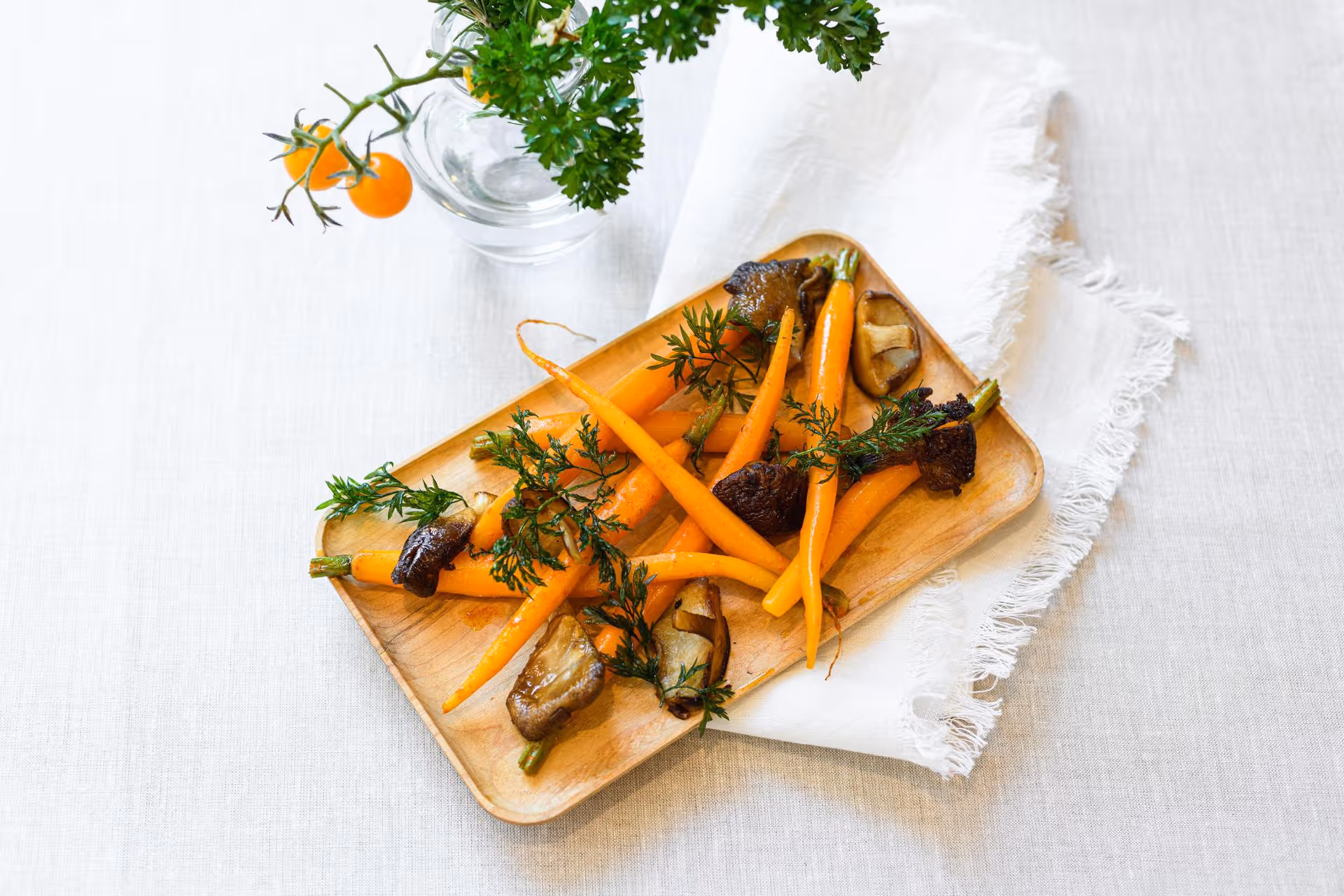 A wooden tray with cooked baby carrots garnished with herbs and mushrooms, placed on a white fringed cloth on a light-colored table. A small glass vase with parsley and two cherry tomatoes is visible in the background.