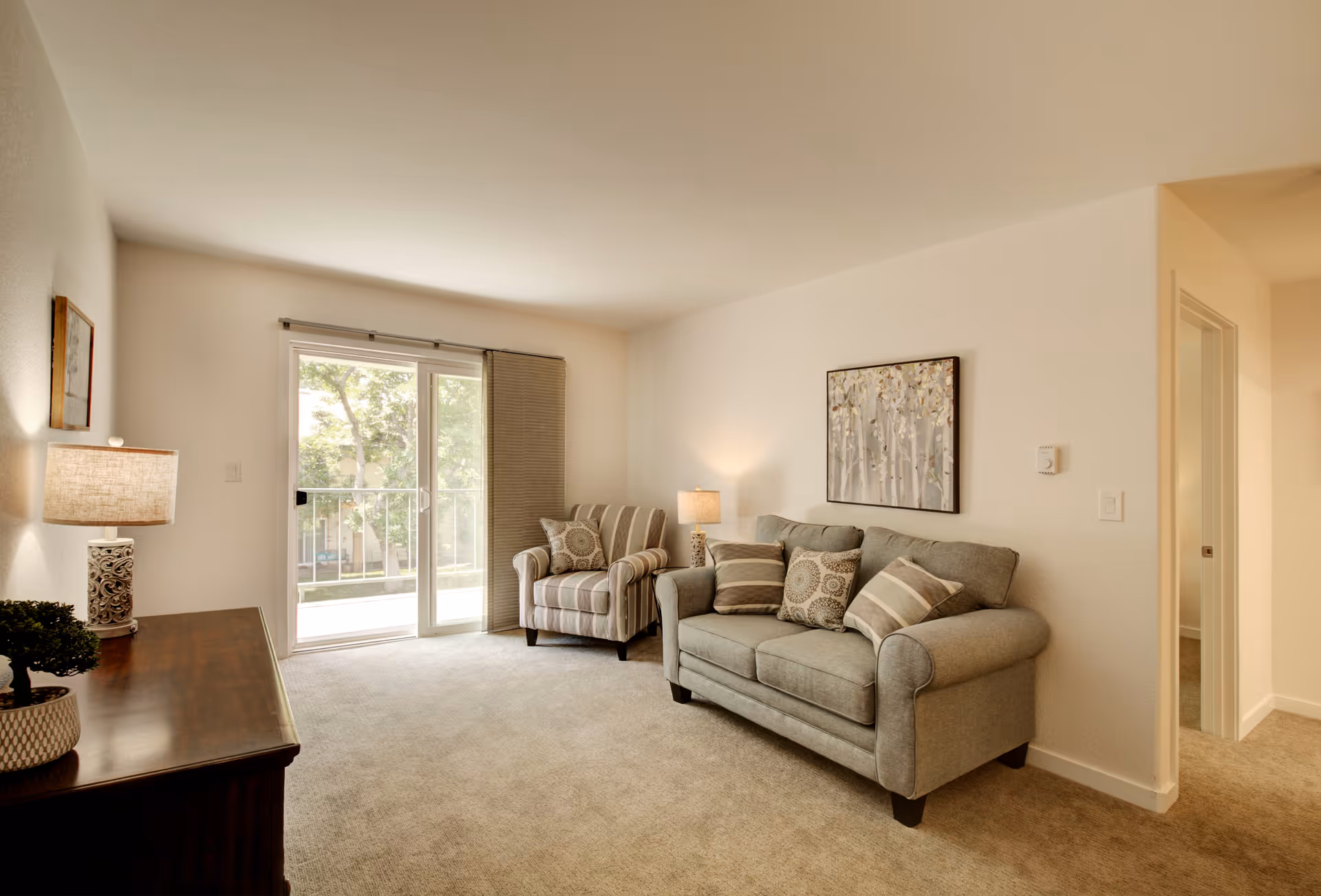 Bright living room with a sofa, armchair, side tables and lamps, wall art, and a sliding glass door to a balcony.