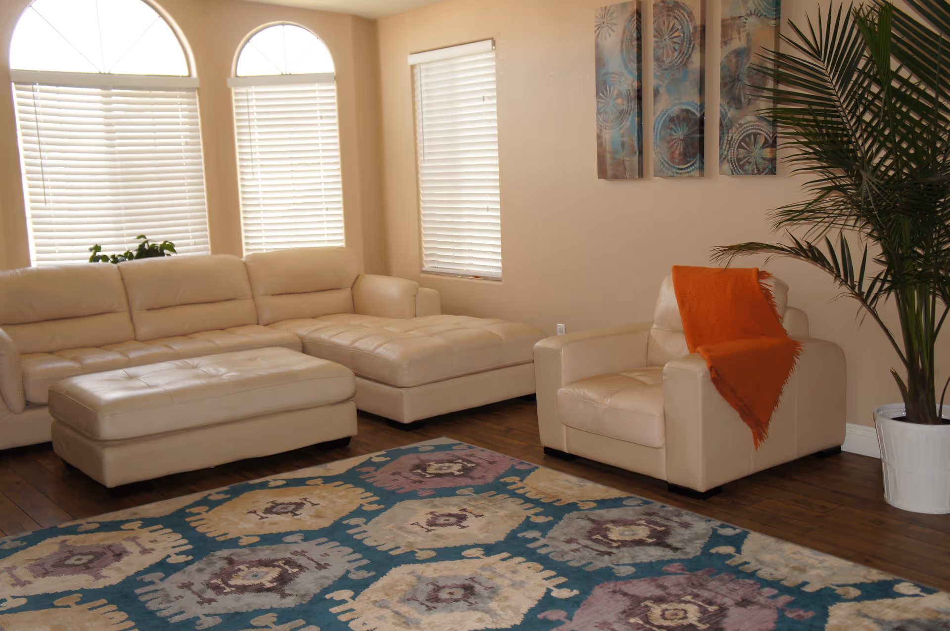 A cozy living room with a beige leather sectional sofa and matching armchair draped with an orange throw blanket. The room features large arched windows with white blinds, a colorful patterned area rug on wooden flooring, a tall green potted plant, and three decorative wall art pieces.