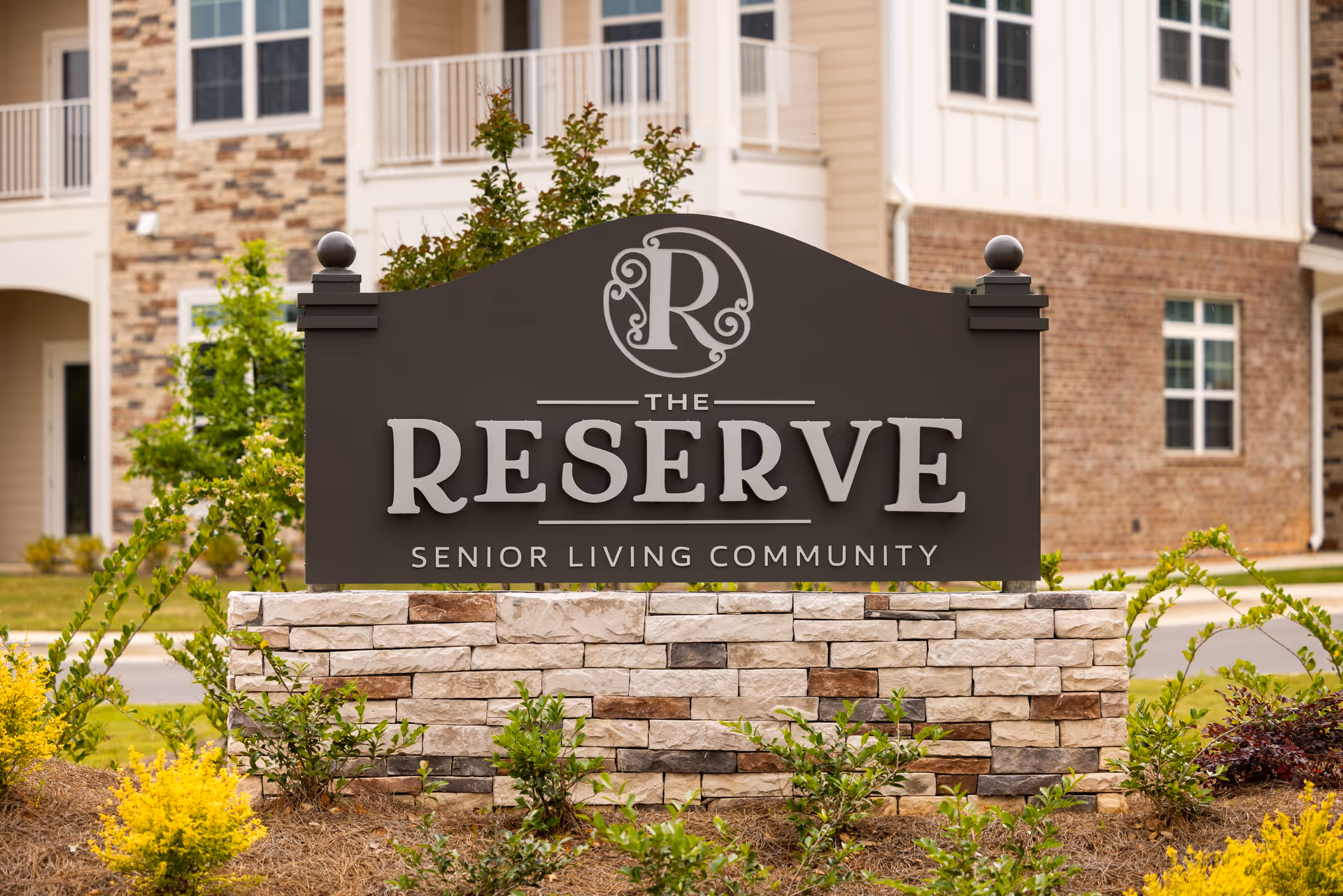 A large sign in front of a senior living community named The Reserve. The sign is dark with white lettering and is mounted on a stone base with landscaping around it. Behind the sign, part of a building with brick and siding is visible.
