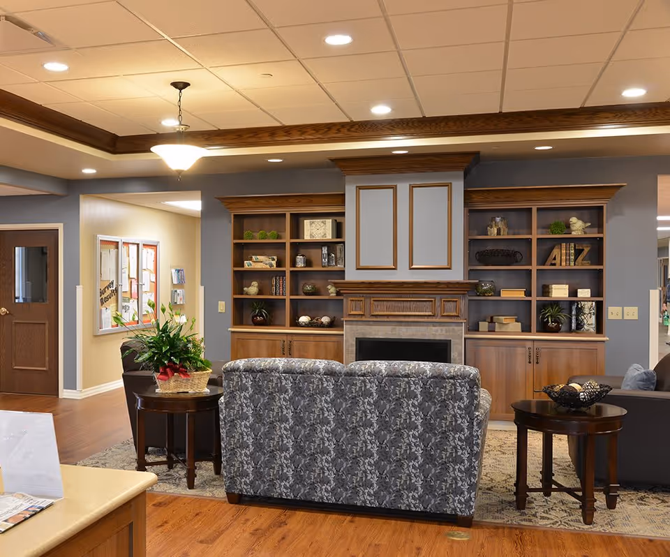 A cozy senior living facility common area with a patterned sofa facing a fireplace surrounded by built-in wooden shelves filled with decorative items. The room has warm wooden flooring, a ceiling light fixture, and a side table with a plant and a basket. There is a bulletin board on the wall near a wooden door in the background.