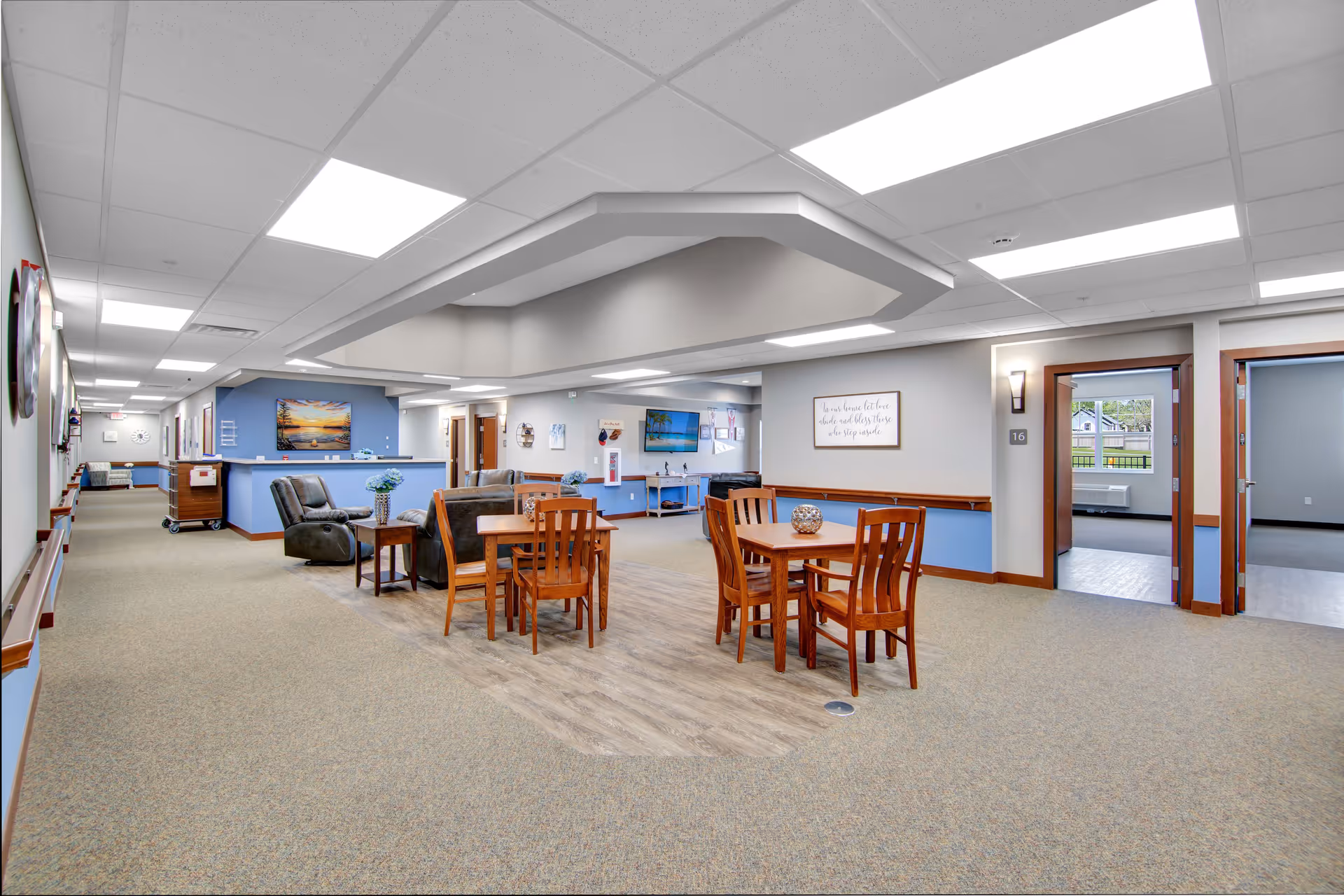 Interior view of a senior living facility common area with carpeted floors and a section of wood flooring. There are two wooden tables each surrounded by four wooden chairs, and two recliner chairs with a small table between them. The walls are painted light gray with blue accents and decorated with framed pictures and a wall-mounted TV. The ceiling has recessed lighting and a unique architectural feature. Doors lead to other rooms, and windows show an outdoor view.