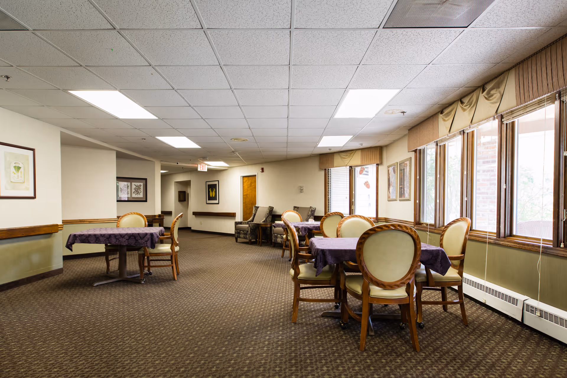 Dining room/common area with round tables covered in purple tablecloths, wooden upholstered chairs, and windows along the right wall.