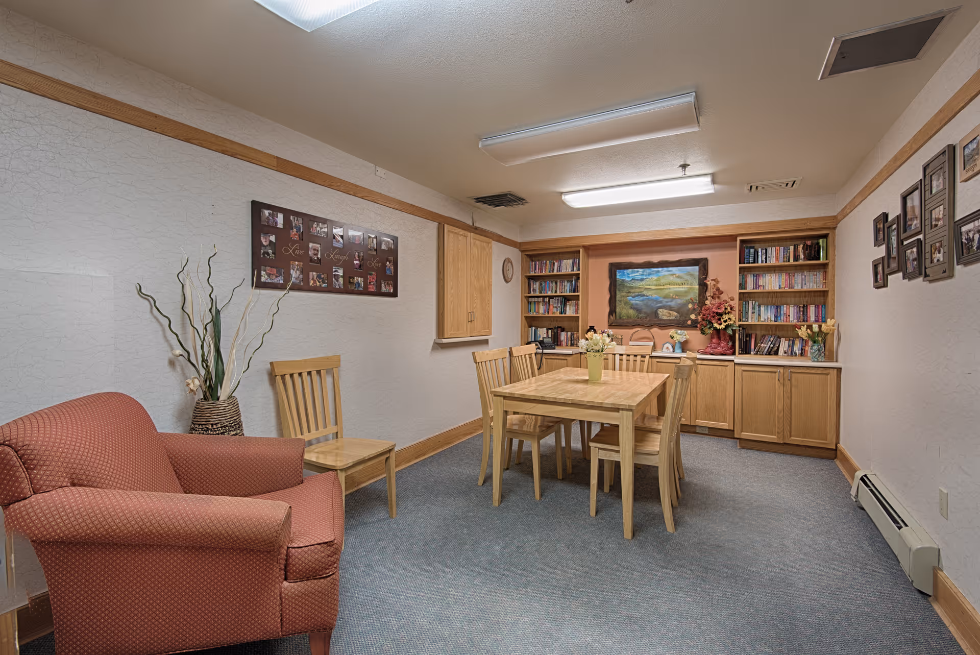 A cozy room with a wooden table and six chairs in the center, surrounded by built-in wooden bookshelves filled with books. There is a red upholstered armchair on the left side, a vase with tall decorative branches next to it, and framed photos on the walls. The room has a carpeted floor and fluorescent ceiling lights.