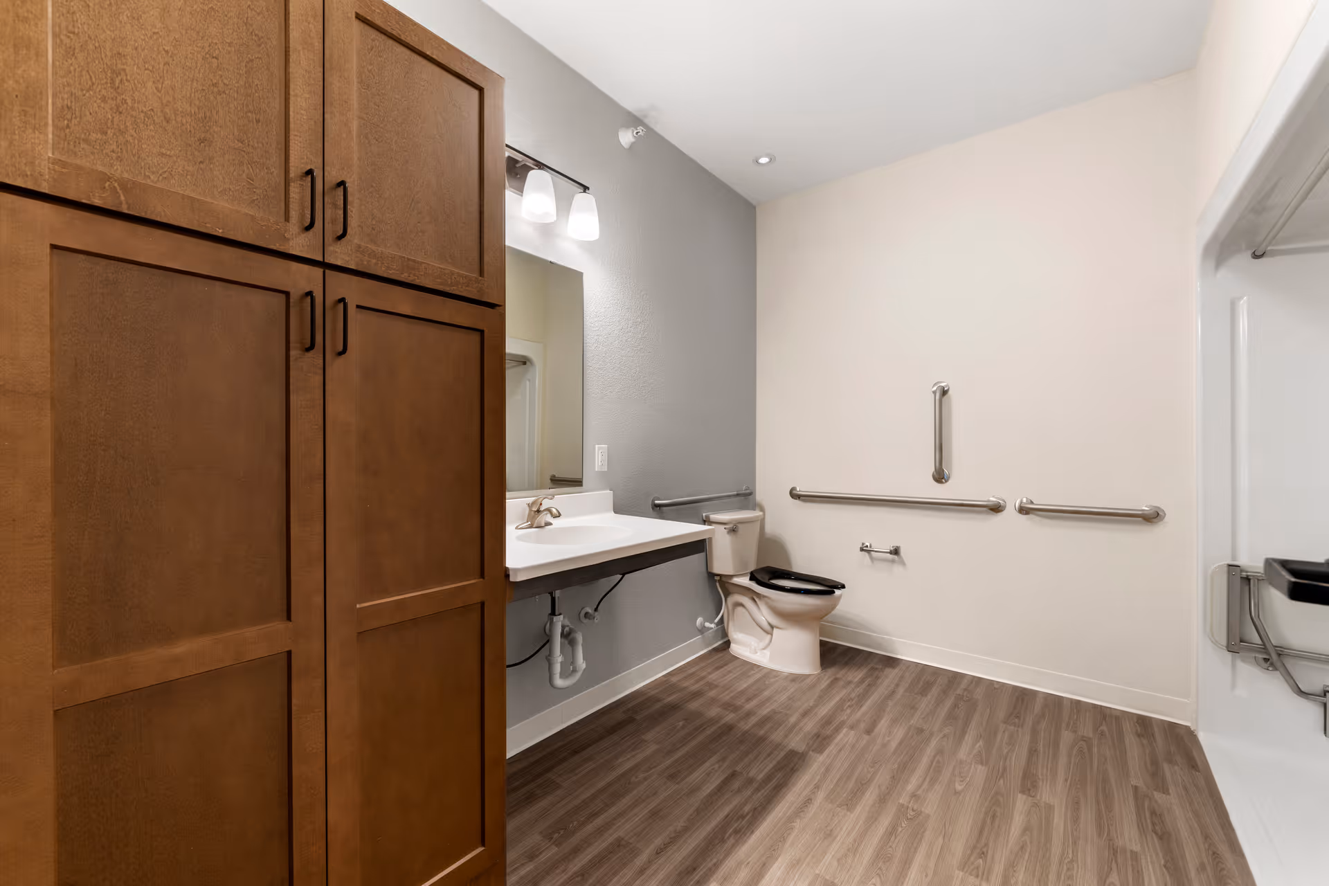 A spacious senior living bathroom with wood-look flooring, a wall-mounted sink with a mirror and light fixture above, a toilet with grab bars on the walls for support, and a walk-in shower with a fold-down seat and grab bars. There is also a tall wooden cabinet for storage.
