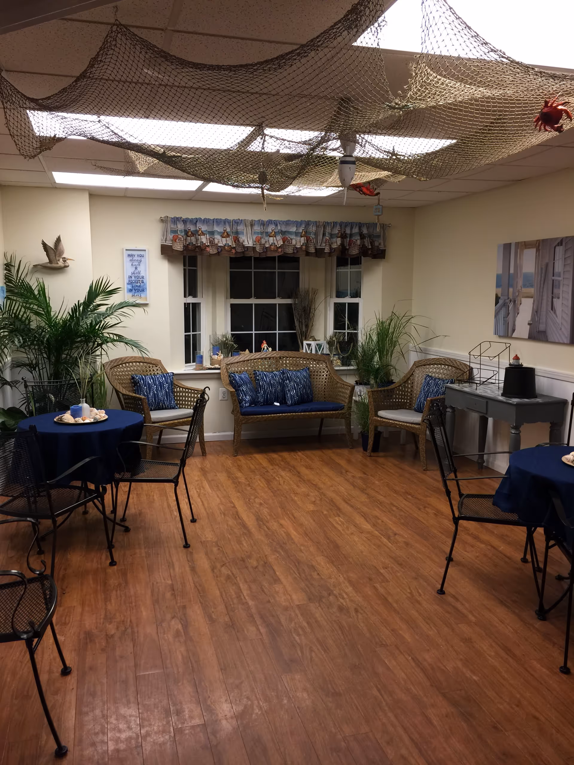 A cozy common area with wooden flooring, wicker chairs with blue cushions, small round tables covered with blue tablecloths, and potted plants. Fishing nets with decorative items hang from the ceiling. A window with a nautical-themed valance is centered on the back wall, and a small gray table with decorative items is on the right side.