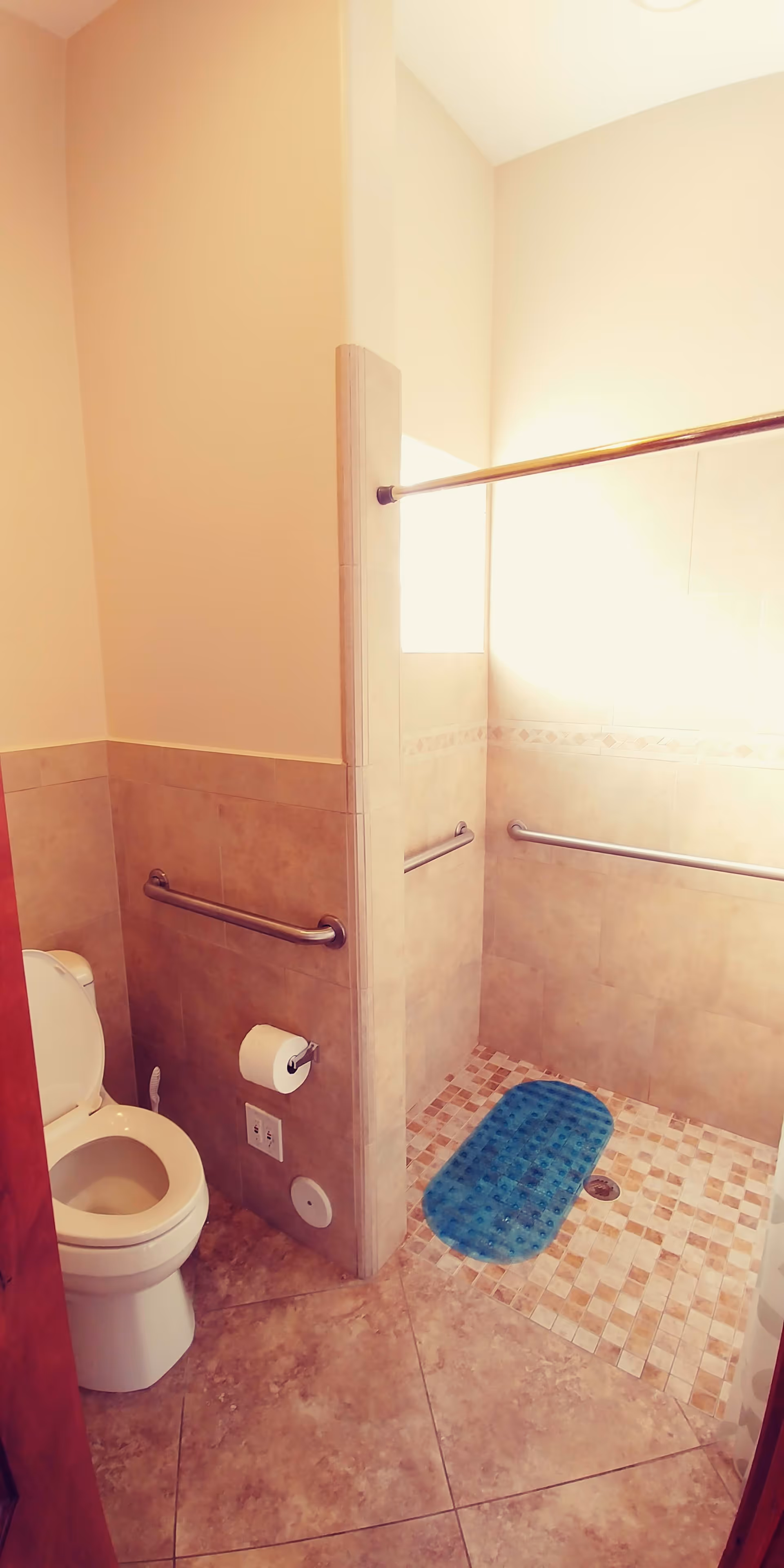 A bathroom with beige tiled walls and floor, featuring a toilet with an open lid on the left side and a walk-in shower area on the right side with a blue non-slip mat on the shower floor. The shower area has grab bars installed on the walls and a metal shower curtain rod above.