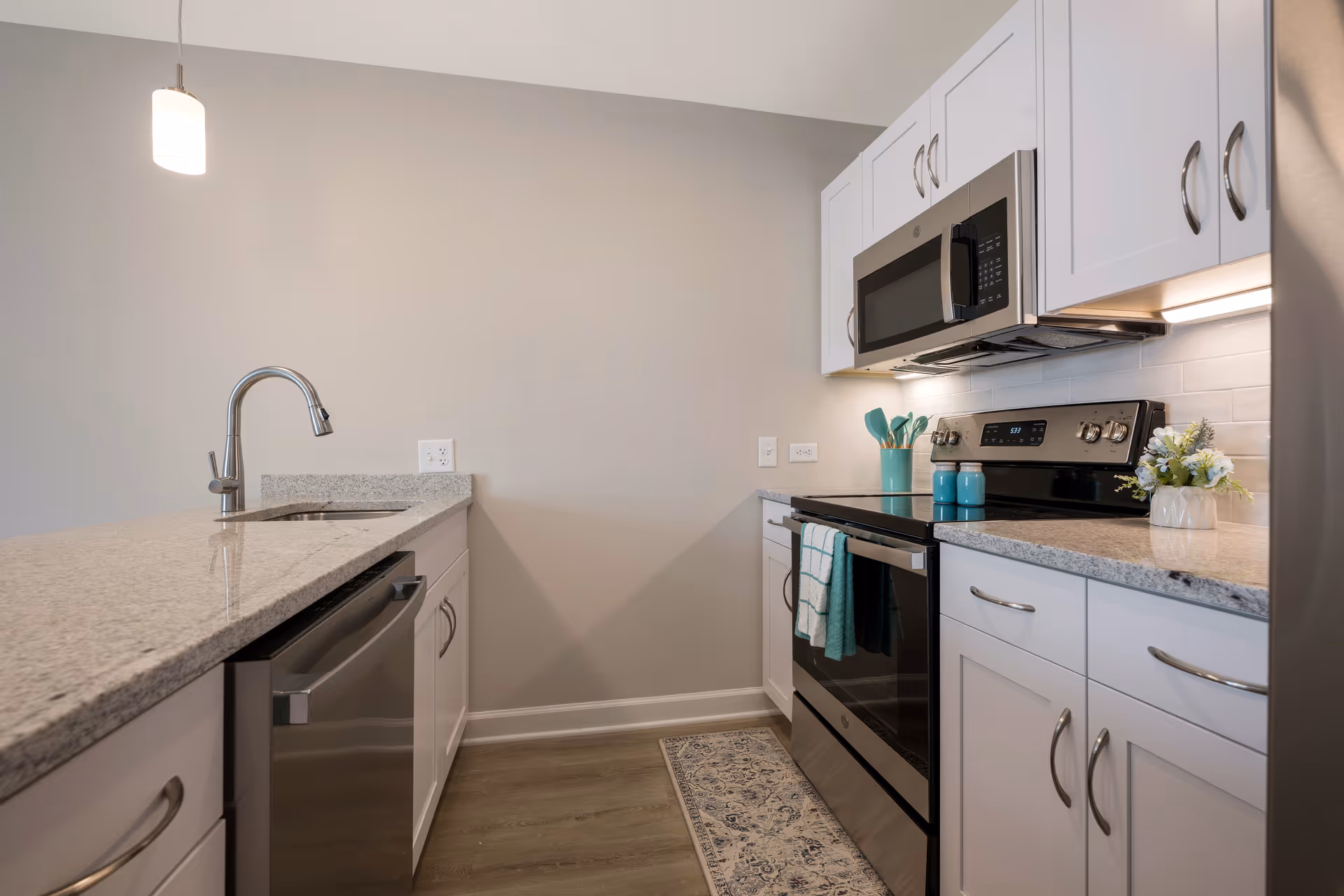 Modern kitchen with white cabinets, granite countertops, stainless steel appliances including a microwave, stove, and dishwasher. There is a sink with a curved faucet on the left countertop, a small pendant light hanging from the ceiling, and decorative items such as a small vase with flowers and kitchen utensils on the right countertop.