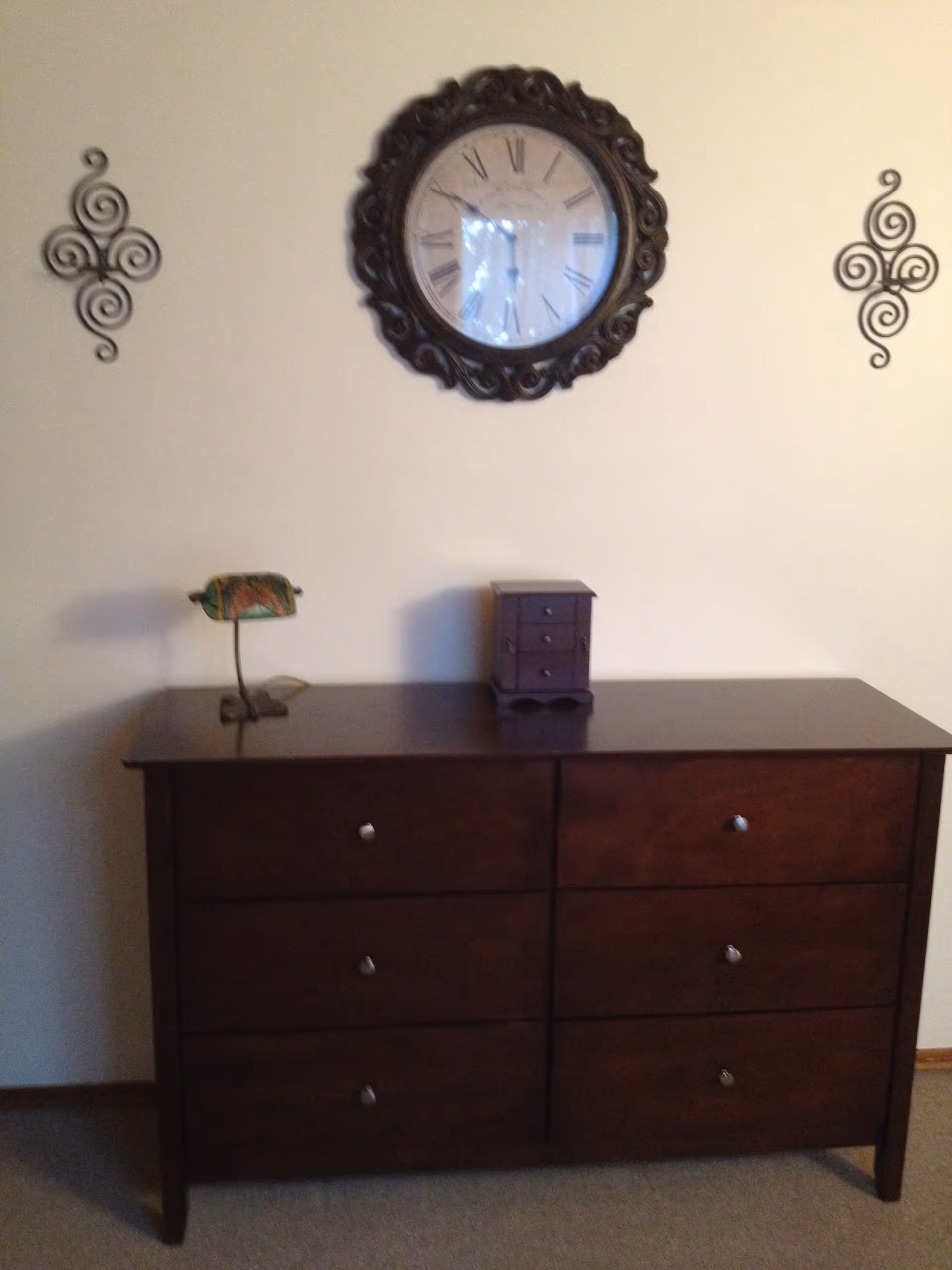 A dark wooden dresser with six drawers stands against a beige wall. On top of the dresser is a small decorative lamp with a stained glass shade and a small wooden jewelry box with three drawers. Above the dresser hangs a large round wall clock with Roman numerals and an ornate dark frame. On either side of the clock are decorative wrought iron wall hangings with spiral designs.