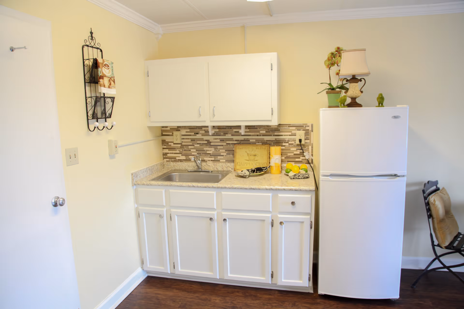 Small kitchen area with white cabinets, a countertop with a sink, a white refrigerator topped with a lamp and a potted plant, and a folding chair with a cushion to the right.