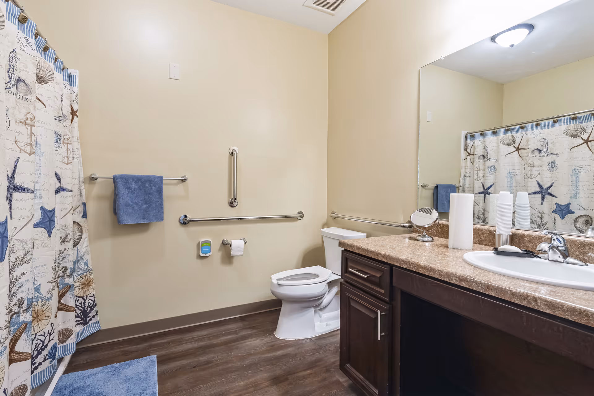 A clean bathroom with beige walls and wood-look flooring. There is a white toilet with grab bars on the walls around it, a blue towel hanging on a towel rack, and a shower with a curtain featuring starfish and seashell designs. A countertop with a sink, paper towels, and a small round mirror is visible, along with a large wall mirror above the sink.