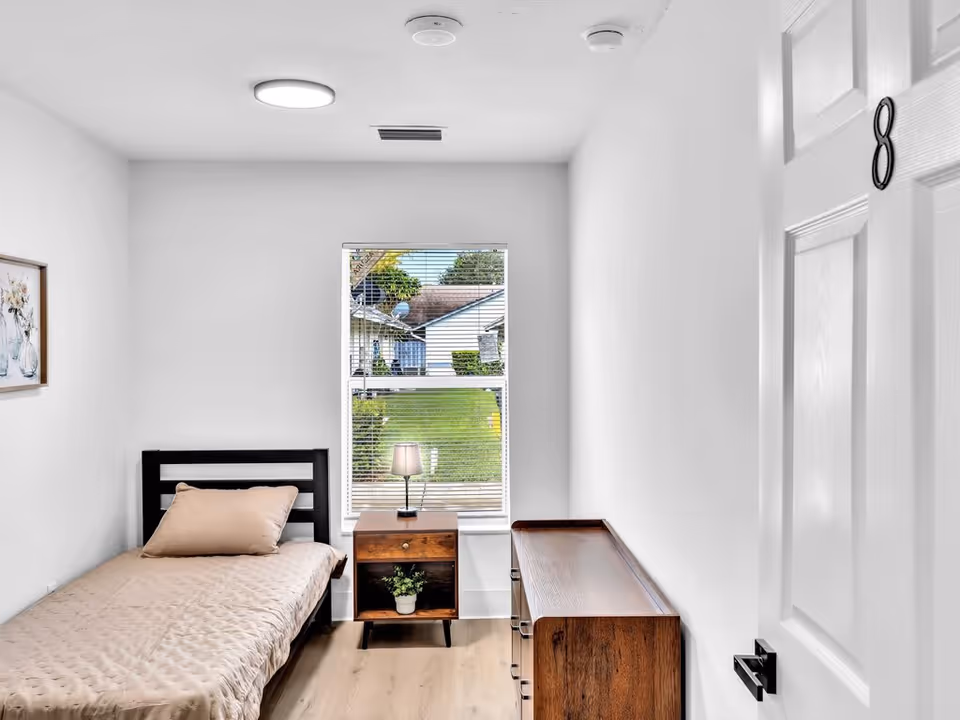 A small, simple bedroom with a single bed covered in a beige quilt and a matching pillow. Next to the bed is a wooden nightstand with a small lamp and a potted plant on the lower shelf. Across from the bed is a wooden dresser. A window with blinds shows a view of a green lawn and neighboring houses. The door to the room is partially open, showing the number 8 on it. The walls and ceiling are white, and the floor is light wood.