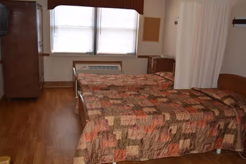A simple bedroom with two twin beds covered in patchwork quilts, a wooden dresser with a TV on top, a window with blinds, and a small air conditioning unit below the window. There is a curtain divider partially visible on the right side of the room.