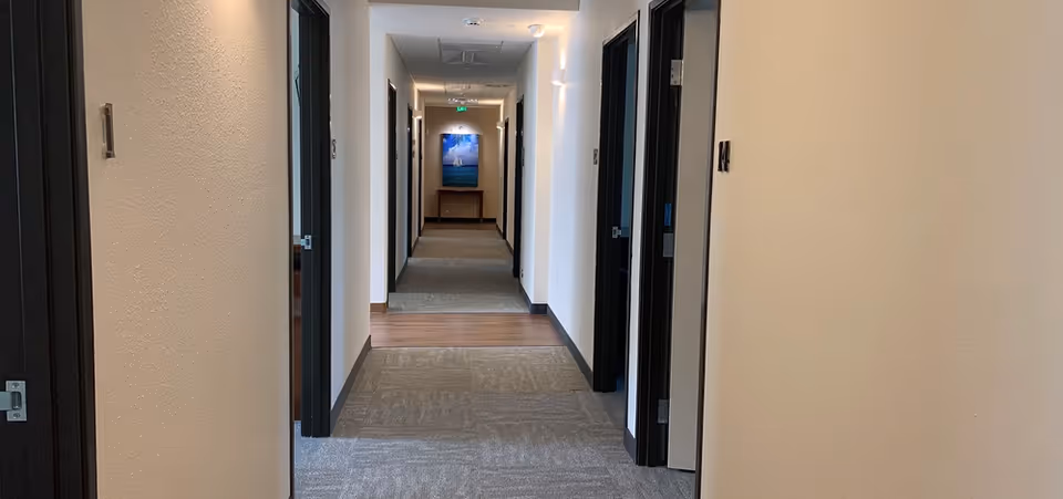 A long, well-lit hallway in a senior care facility with multiple doorways on both sides. The floor is carpeted with a section of wood flooring near the end. A piece of artwork and a small table are visible at the far end of the hallway.
