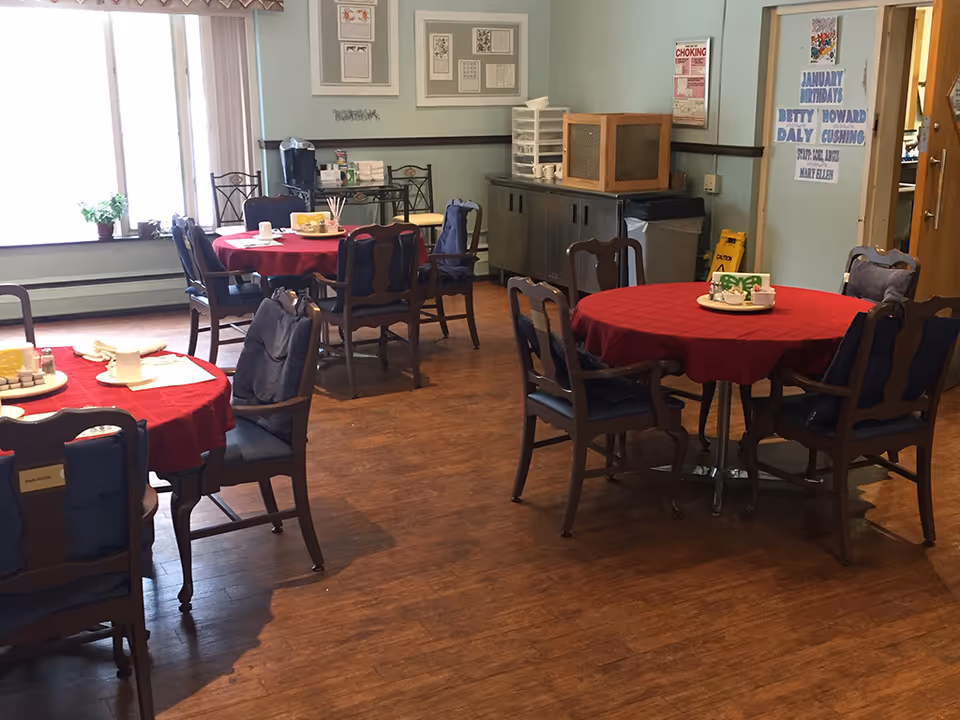 A dining room with several round tables covered with red tablecloths and surrounded by wooden chairs with blue cushions. The room has wooden flooring, a large window with potted plants on the windowsill, and a sideboard with various items including a coffee maker. There are framed pictures on the wall and a poster listing January birthdays.