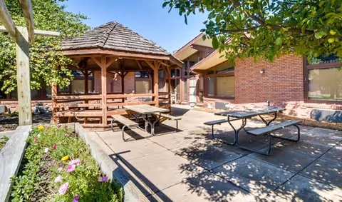 Outdoor patio area at Cobblestone Court featuring a wooden gazebo with a shingled roof, picnic tables on a concrete surface, surrounded by trees and flower beds, adjacent to a brick building with large windows.