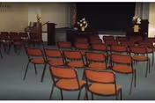 Interior meeting/chapel room with rows of orange chairs facing a small stage with podiums and floral arrangements.