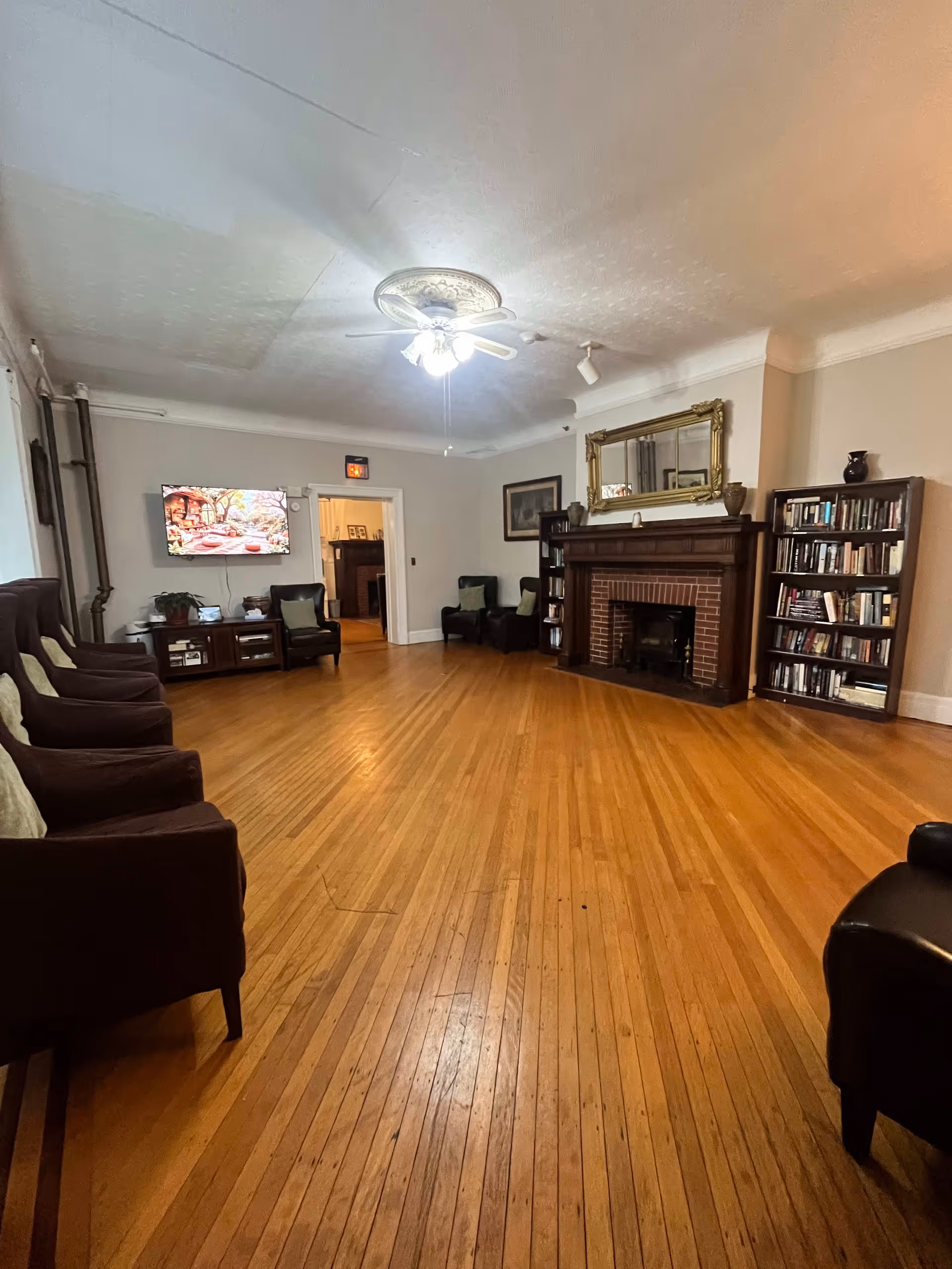 A spacious living room with wooden floors, several dark upholstered chairs arranged along the walls, a brick fireplace with a large ornate gold-framed mirror above it, bookshelves filled with books on either side of the fireplace, a ceiling fan with lights, and a flat-screen TV mounted on the wall displaying a nature scene.