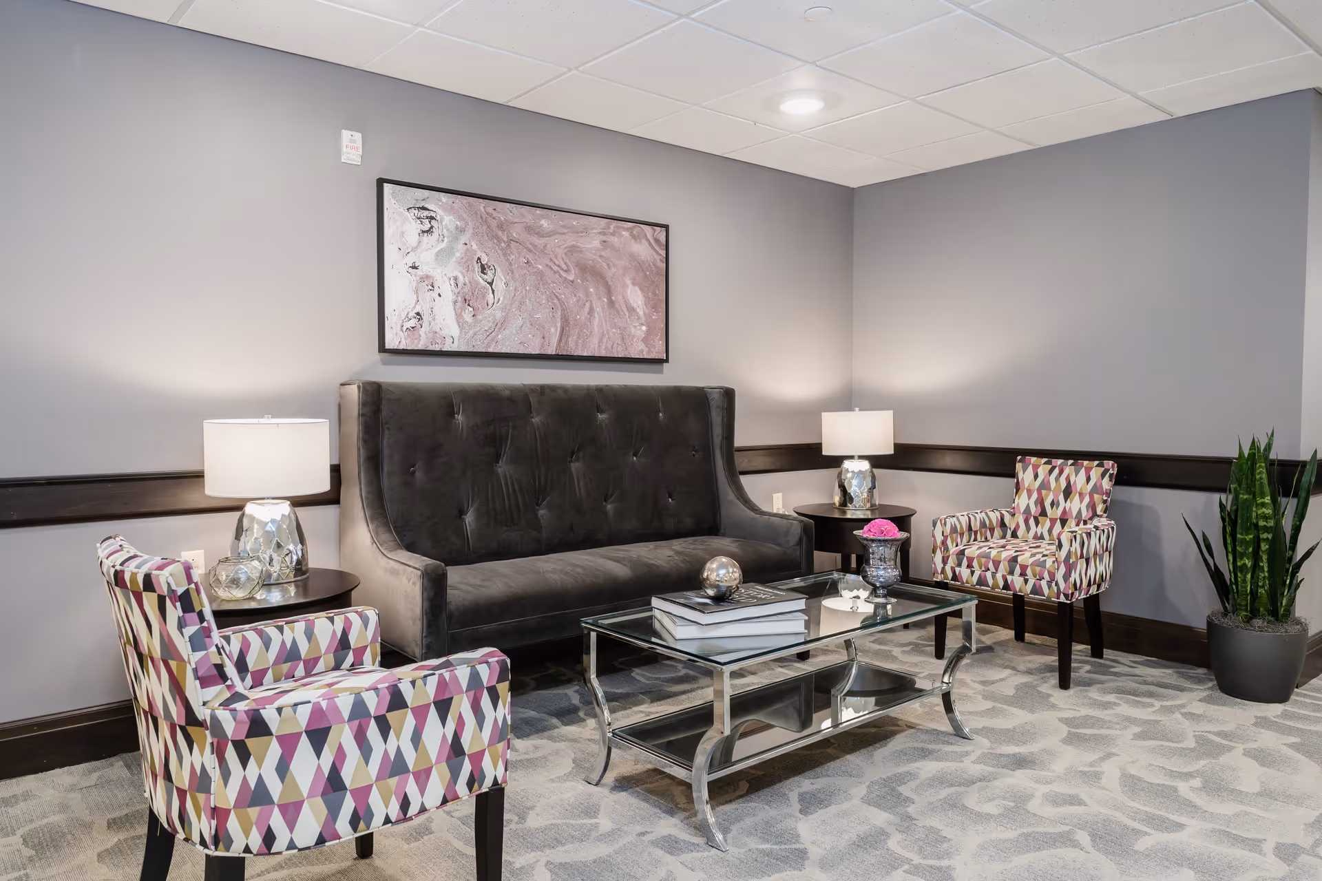 A modern sitting area with a dark gray tufted sofa, two patterned armchairs in shades of purple, pink, and beige, a glass coffee table with books and decorative items, two side tables with lamps, a large abstract artwork on the wall, and a potted plant in the corner.