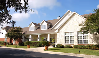 Exterior view of a senior living facility building with a stone facade, large windows, a covered porch with white columns, and a well-maintained lawn with potted plants and trees under a blue sky with some clouds.