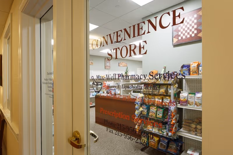 View through a glass door into a convenience store with shelves of snacks and a prescription counter, the words 'CONVENIENCE STORE' visible on the glass.