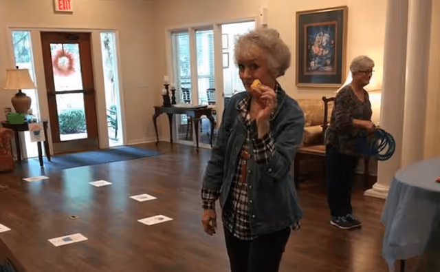 Two elderly women in a senior living facility common area. One woman in the foreground is holding a small yellow object and looking at the camera. The other woman in the background is standing near a column and holding several blue rings. The room has wooden floors, a table with a lamp, framed artwork on the wall, and a door with glass panels leading outside.