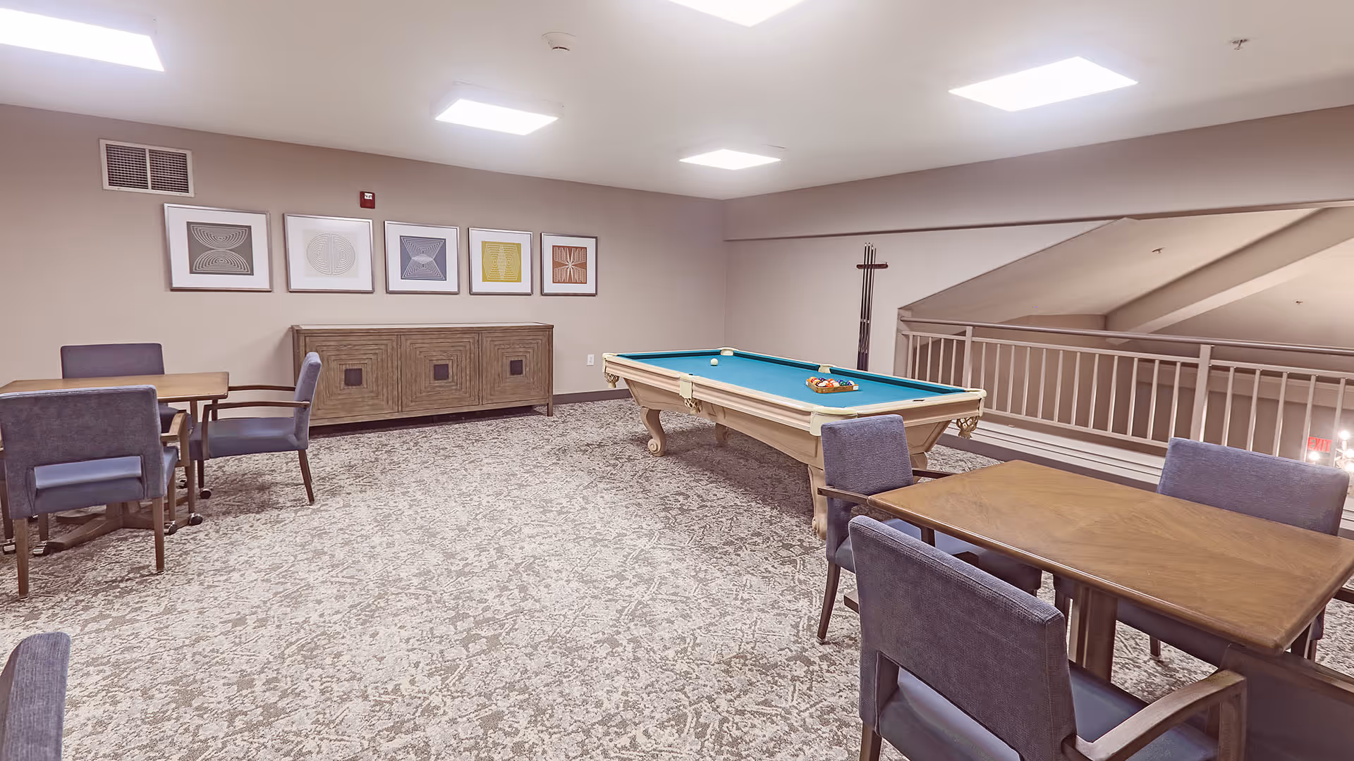 A recreational room with a pool table in the center, surrounded by several wooden tables and chairs with blue upholstery. The room has beige walls, a patterned carpet, and five framed abstract artworks hanging on the wall above a wooden sideboard. There is a railing on the right side overlooking a lower level.