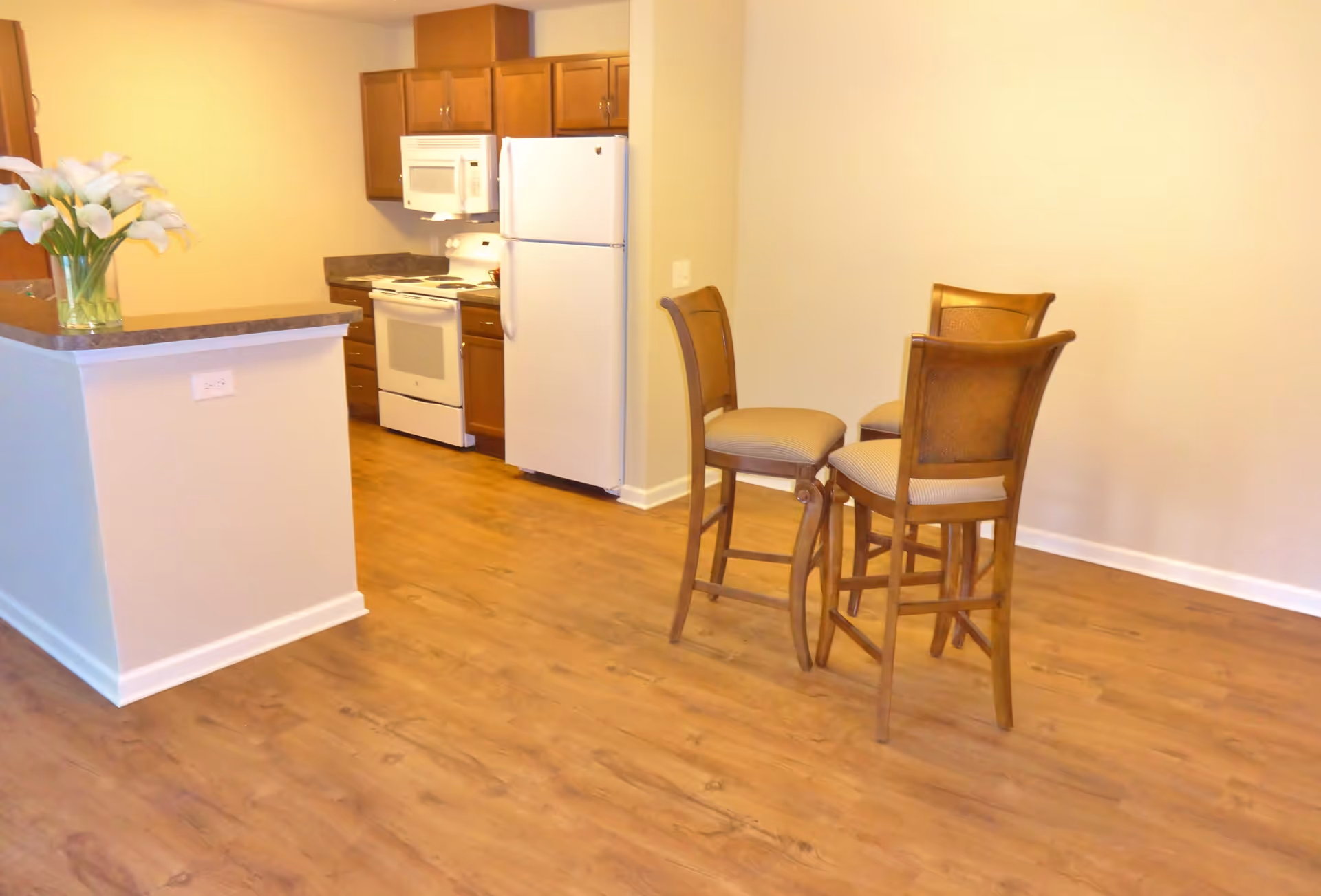 A kitchen area with wooden cabinets, a white refrigerator, stove, and microwave. In the foreground, there is a small dining area with three wooden chairs with cushioned seats. A vase with white flowers is placed on the kitchen counter. The floor is wooden, and the walls are painted a light beige color.