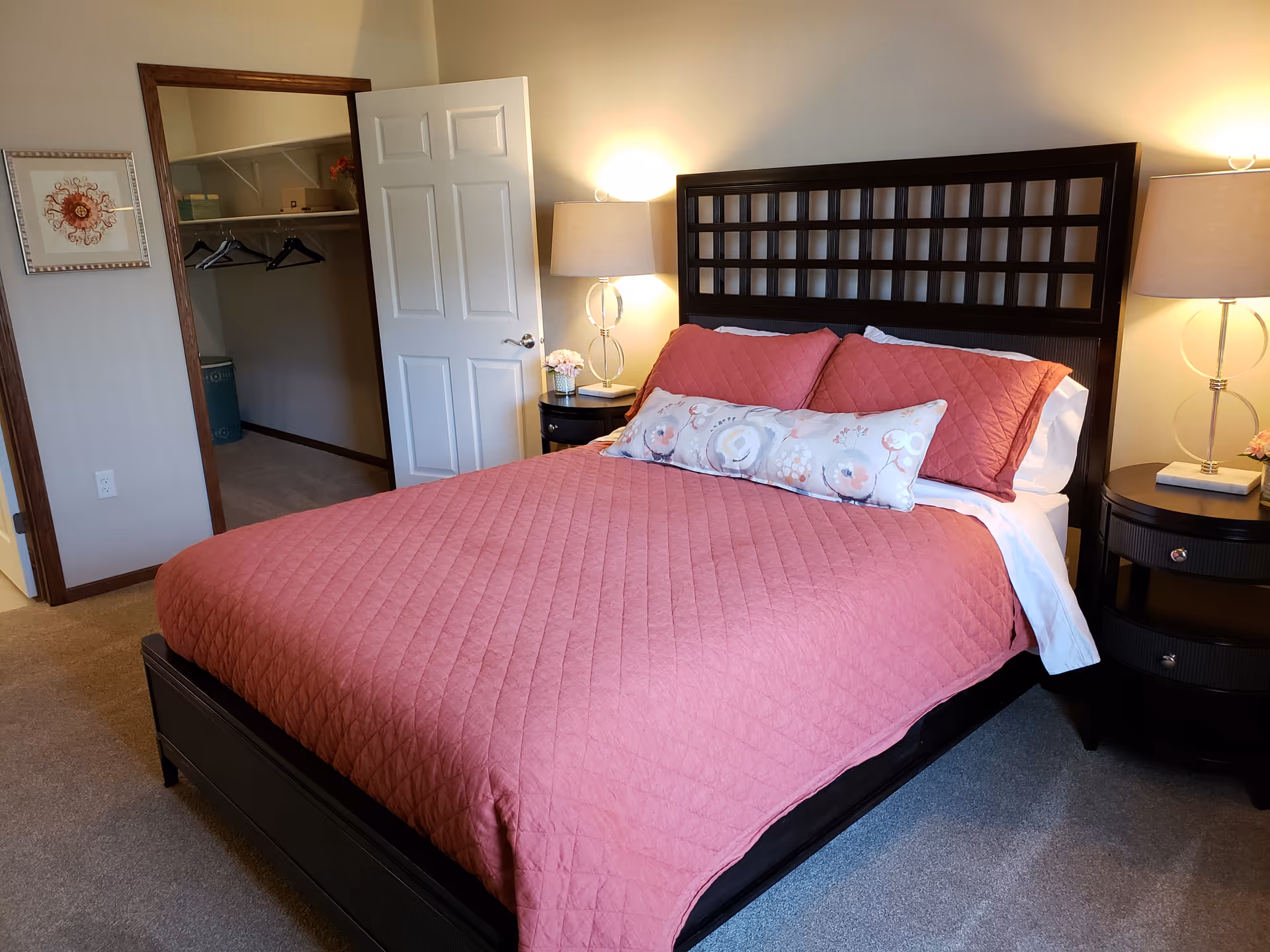 A neatly made bedroom with a pink quilted bed, two bedside tables with lamps, and an open closet door.