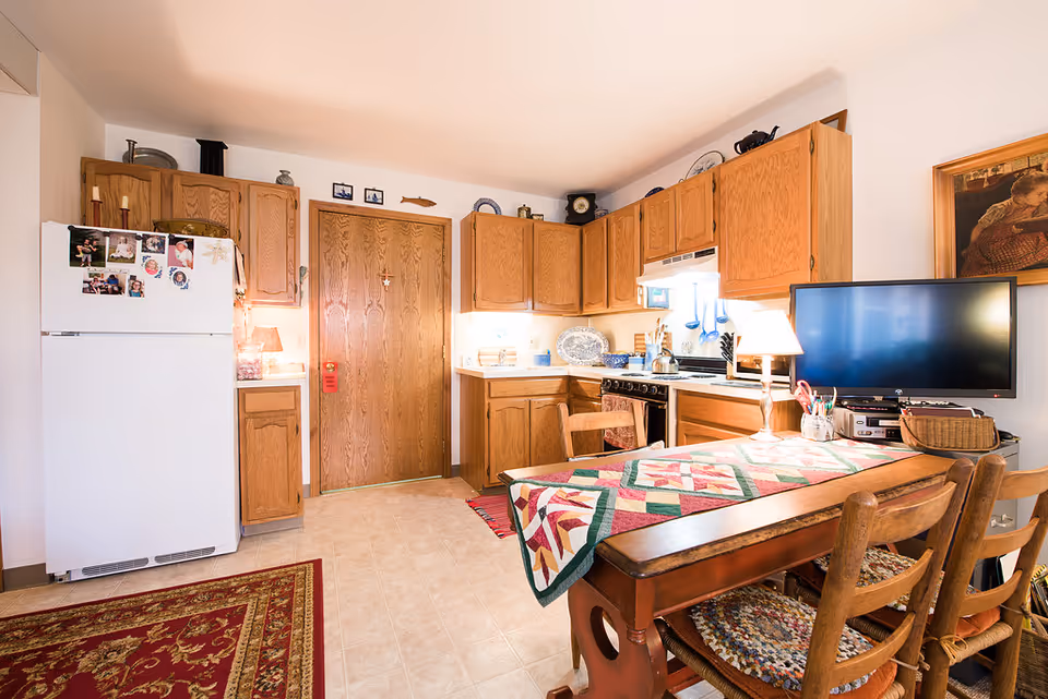 A cozy kitchen with wooden cabinets, a white refrigerator adorned with family photos and magnets, a stove with a kettle, and a wooden dining table covered with a colorful quilted table runner. The room is warmly lit with a table lamp and natural light coming through a window. There is a framed painting on the wall and a red patterned rug on the floor.