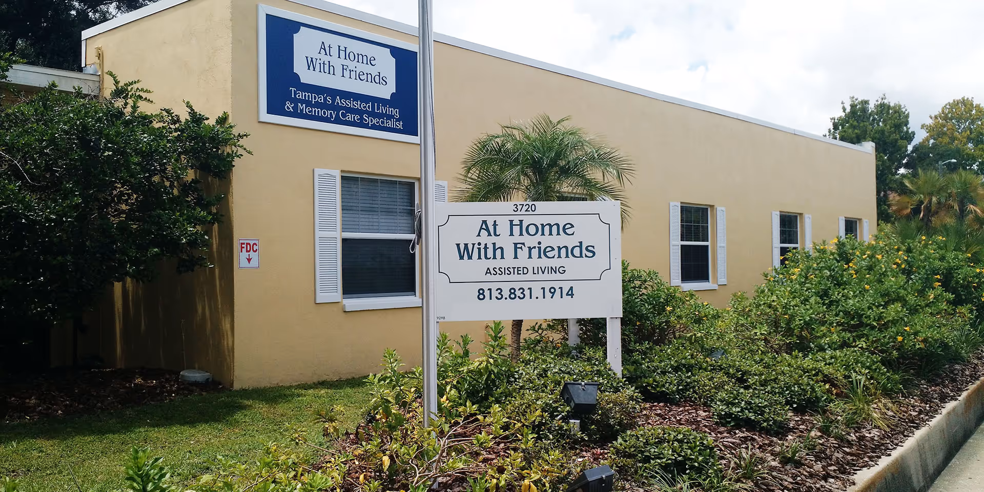 Beige single-story assisted living building with a white sign reading 'At Home With Friends' in a landscaped front yard.